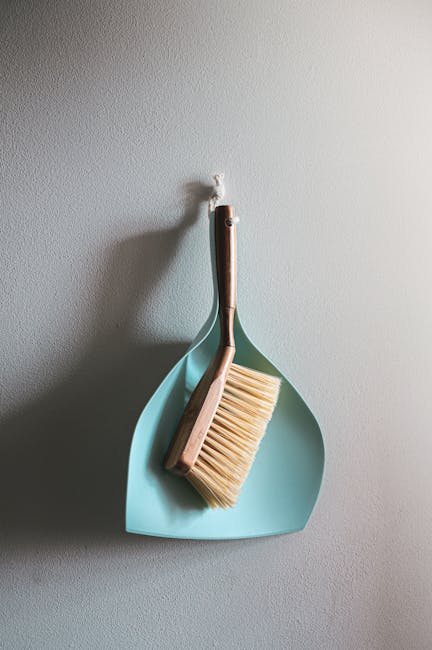 A light blue dustpan hangs on a plain white wall, with a vegetable brush and a scrub brush resting inside it. The brushes have wooden handles and bristles, showing signs of use. The scene is well-lit,