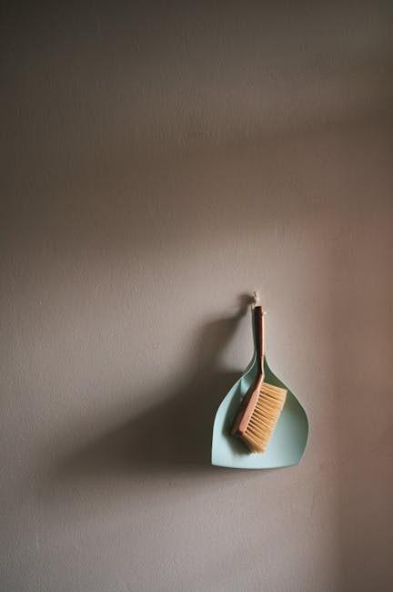 A light blue dustpan hangs on a plain white wall, with a vegetable brush and a scrub brush resting inside it. The brushes have wooden handles and bristles, showing signs of use. The scene is well-lit,